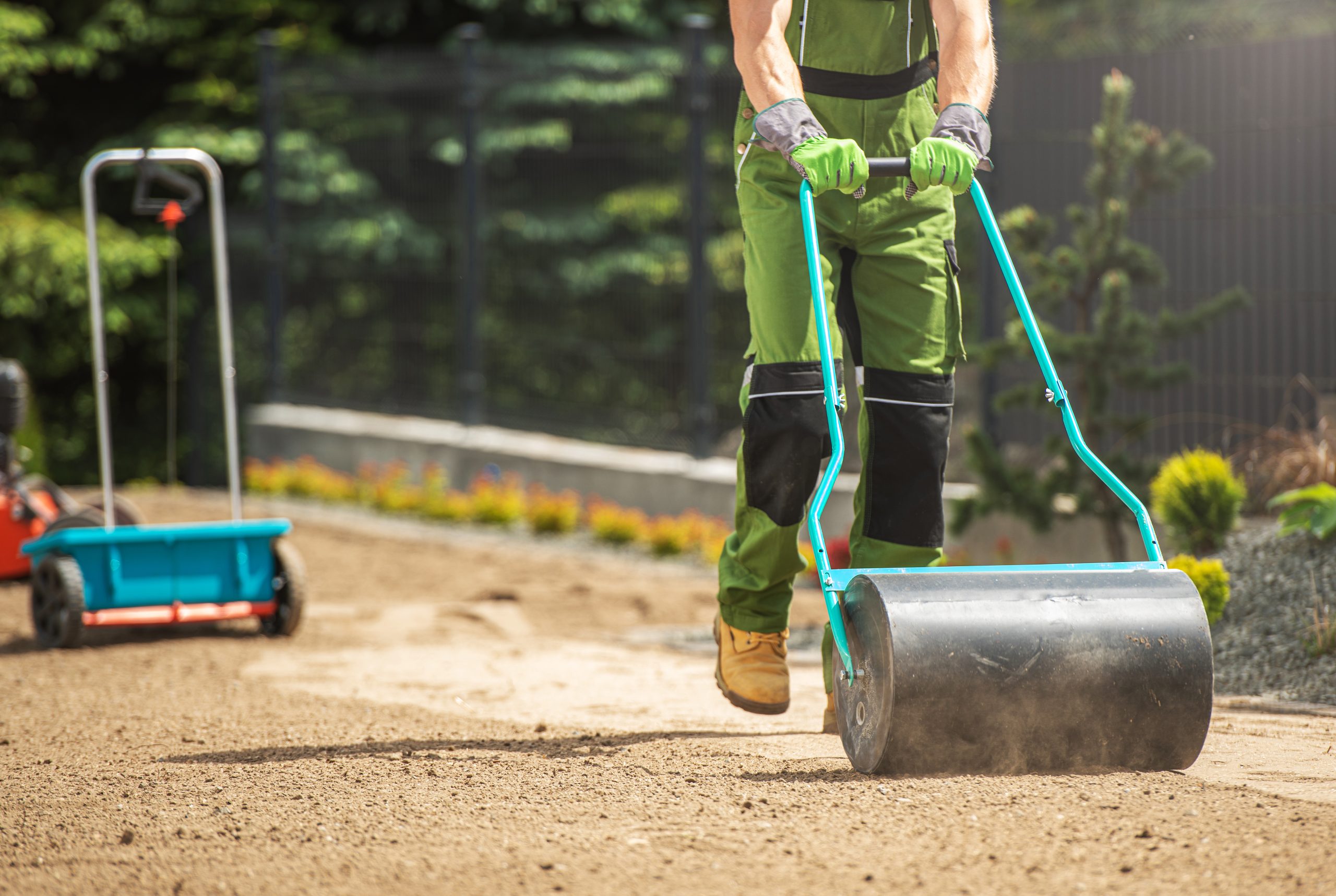 Poly Push Tow Lawn Roller Pushed by Caucasian Gardener. Soil Compacting Before Grass Seeding. Preparing Garden Landscape. Gardening and Landscaping Theme.